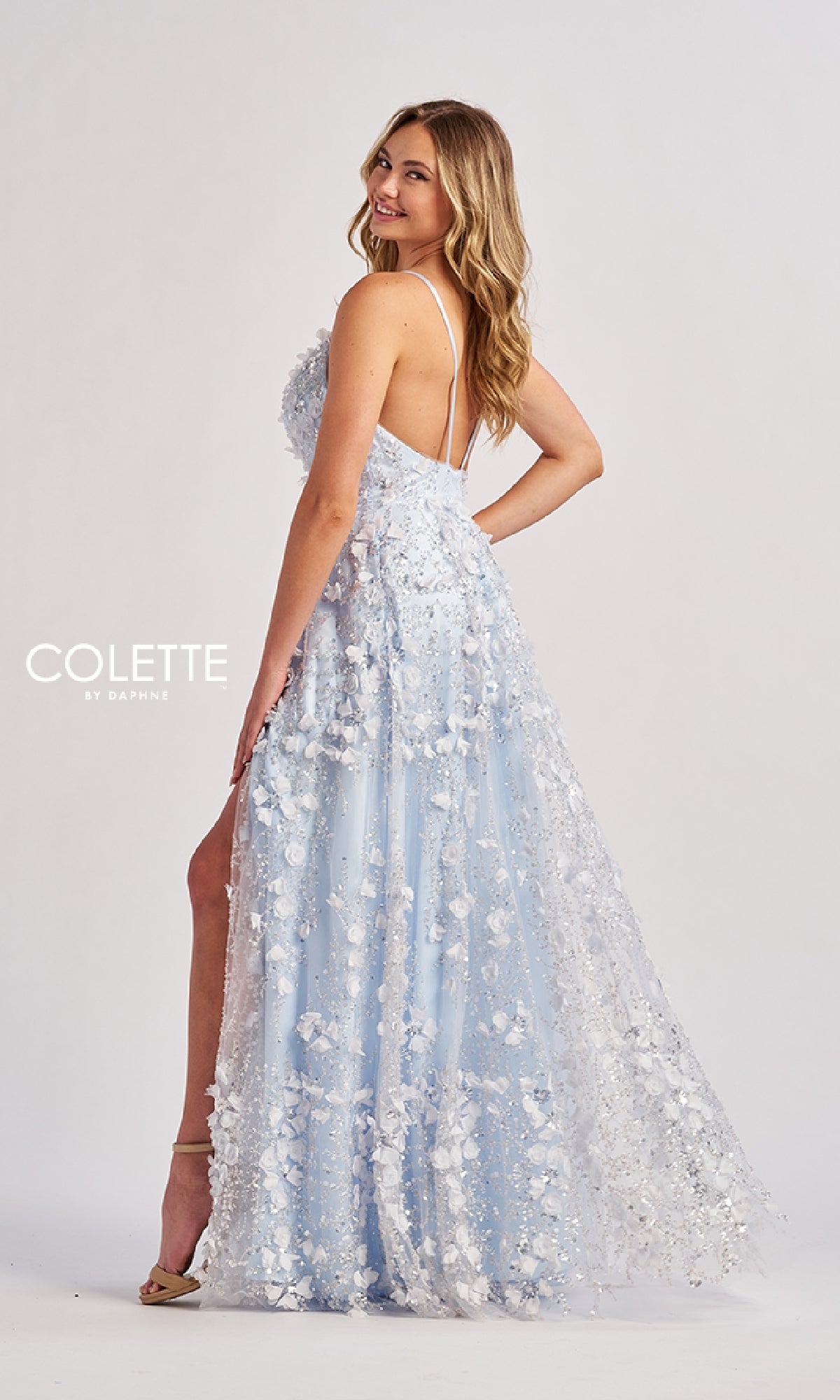 A woman in the Colette CL8525 long prom dress—a light blue, floor-length A-line gown with spaghetti straps and flower embellishments—stands sideways, smiling against a plain background.